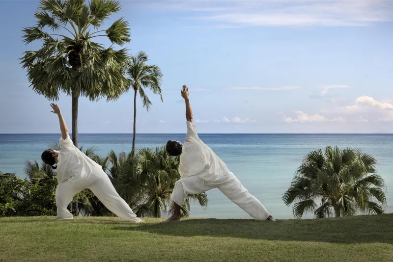 Two guests in white doing a yoga triangle pose on a lawn above the sea at Anantara Lawana, Koh Samui