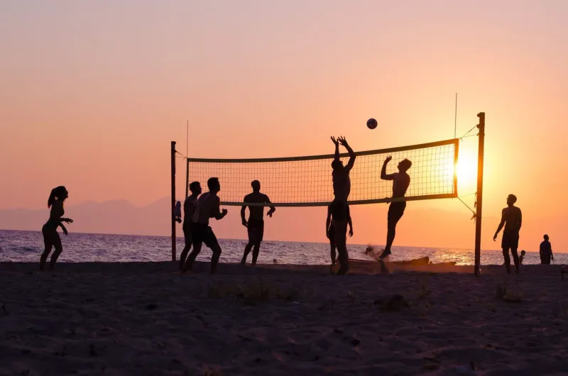 Silhouetted players set and spike a volleyball over a net on the beach as the sun sets over the water