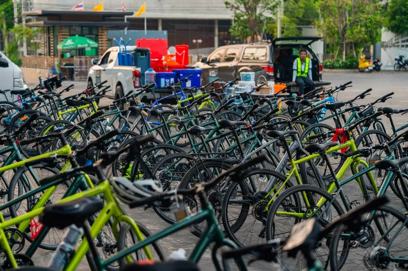 Rows of green and lime hybrid rental bicycles parked in a Thai car park beside support trucks