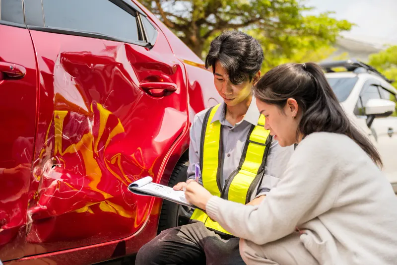Two people inspecting a dented red car door and writing on a clipboard