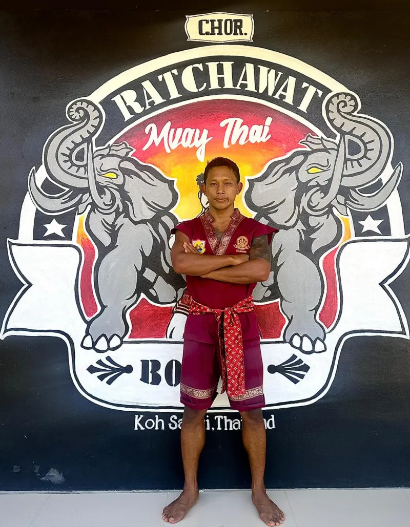 A fighter in a red ceremonial robe standing in front of the elephant logo on the gym wall
