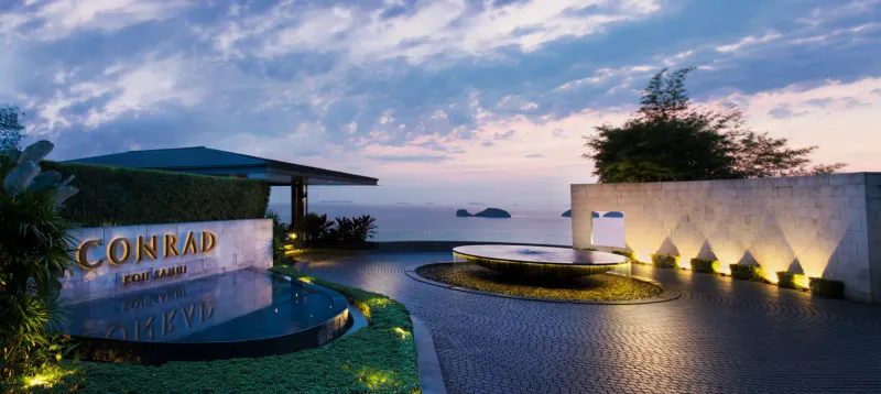 Conrad Koh Samui entrance at dusk with lit signage, a round fountain and the Five Islands beyond