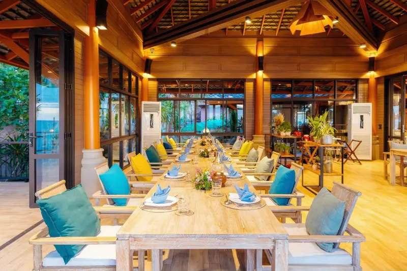 Long set dining table with colourful cushions inside a teak-beamed open restaurant pavilion
