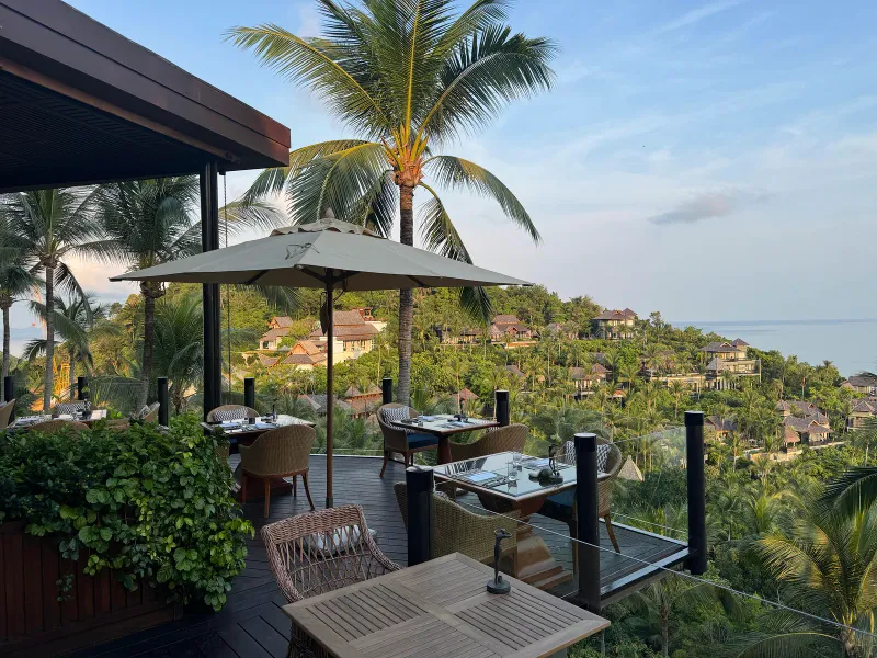 Open-air restaurant terrace with tables, umbrella and palms above the hillside villas at Four Seasons Resort Koh Samui