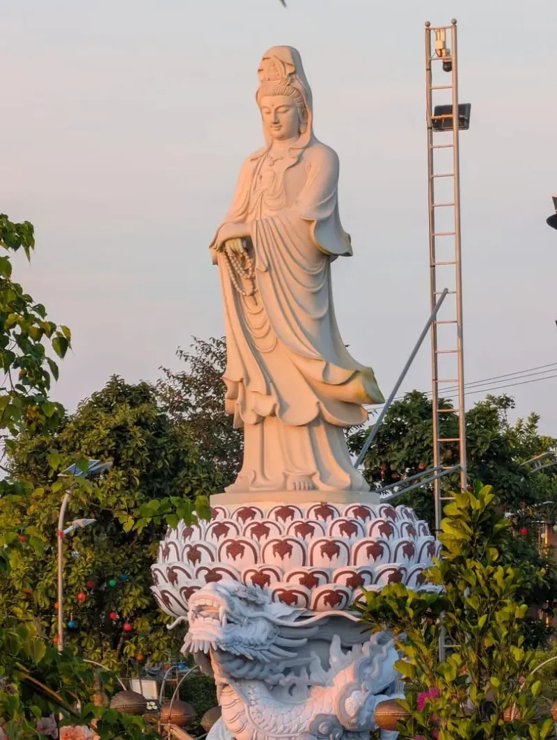 A peaceful white statue of Guanyin standing on a dragon pedestal amidst lush greenery outdoors