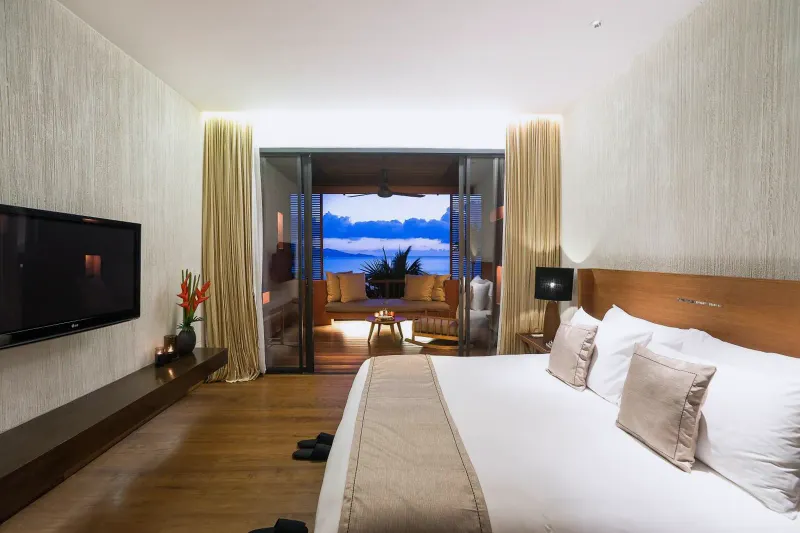 Bedroom at Hansar Samui with a large bed, television and a glimpse of the sea through the balcony doors