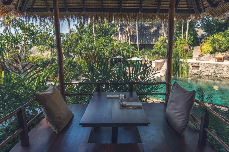 An open-air dining pavilion table set beside a turquoise pool in the jungle