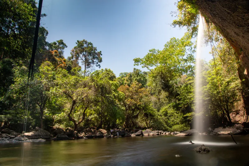 A jungle waterfall dropping into a calm river pool beneath an overhanging rock ledge on Koh Samui
