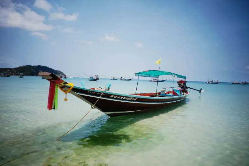 Colourful wooden longtail boat moored in clear shallow water with fishing boats behind