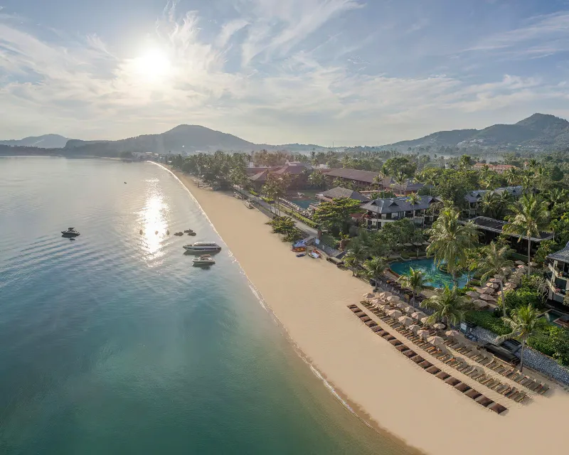Aerial view of a wide Koh Samui bay with a sandy beach, palm-lined resorts and boats on calm water