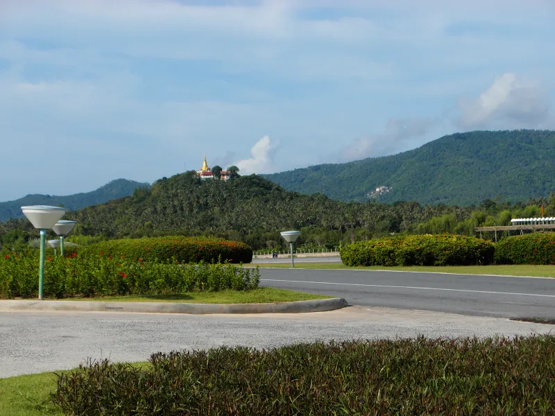 Road and landscaped verge outside Koh Samui Airport with forested hills behind