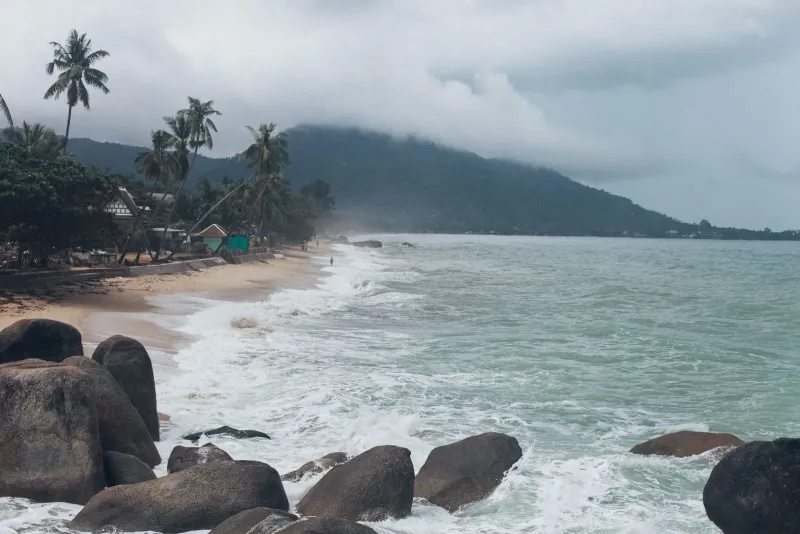 Waves crashing on a Koh Samui beach with lush green hillside, typical of early December's leftover monsoon swell
