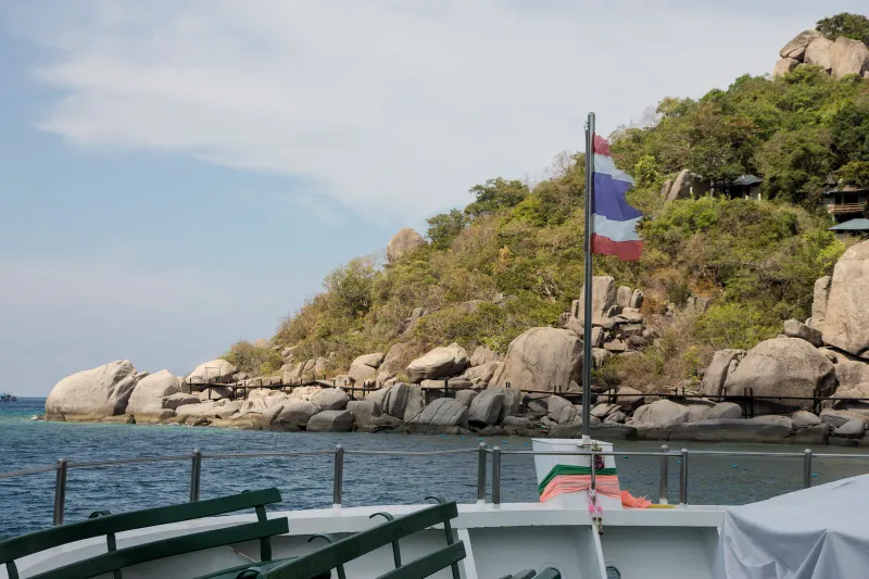 View from a boat deck of a Thai flag and a boulder-strewn forested island shore