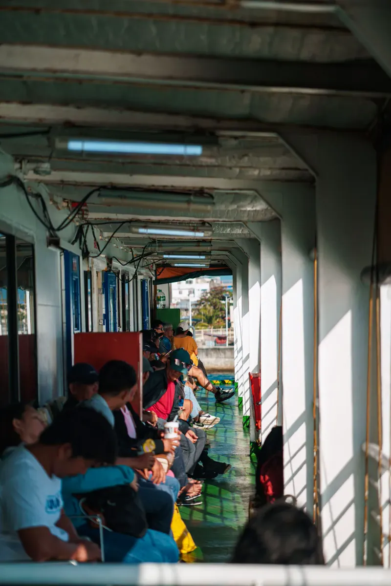 Passengers seated along the shaded side deck of a ferry in the afternoon sun