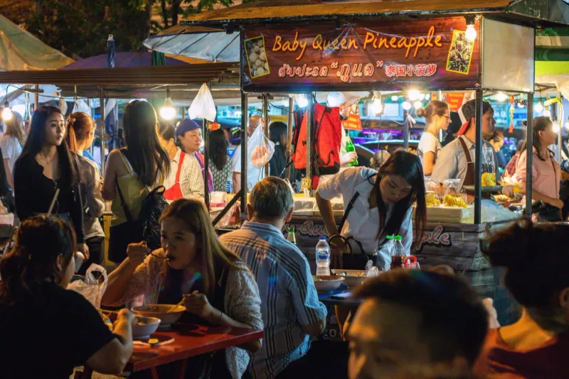 Crowded night market street with colorful food stalls and shoppers browsing under string lights