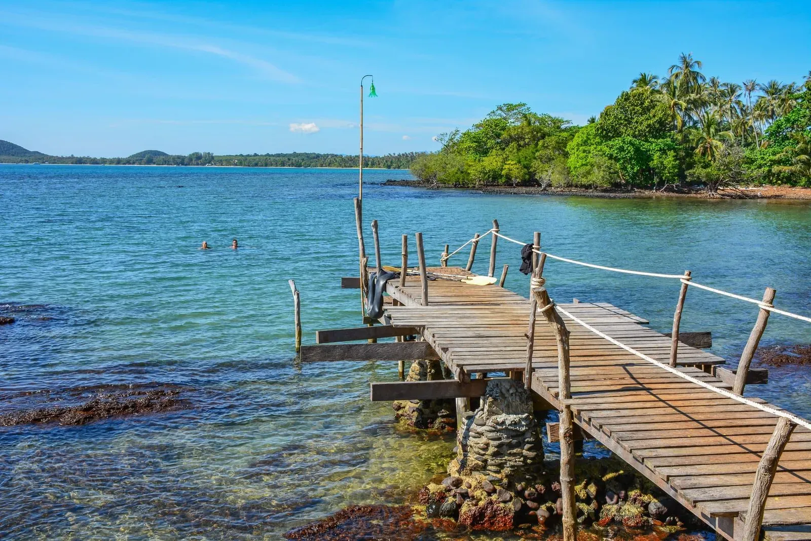 Rustic wooden jetty with rope rails reaching over clear turquoise water and dark reef rocks at Koh Madsum, a palm-covered point on the right, two swimmers in the bay
