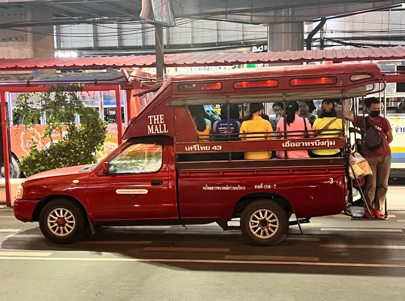 Red songthaew pickup truck with passengers on its benches at a Thai town stop
