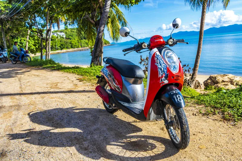 Red Honda scooter parked on a sandy track beside a quiet bay on Koh Samui