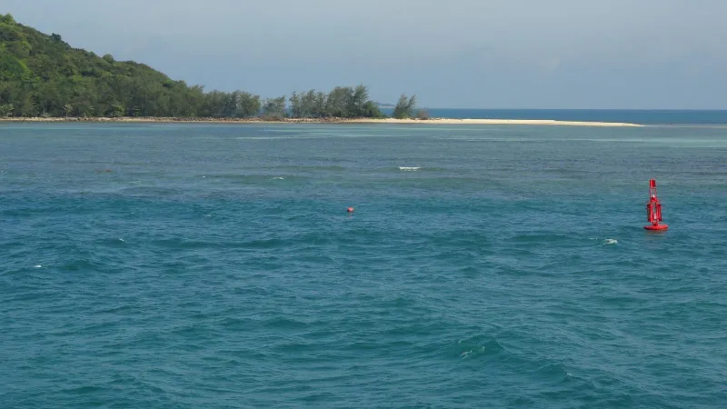 Red navigation buoy in choppy blue water off a forested island in the Gulf of Thailand