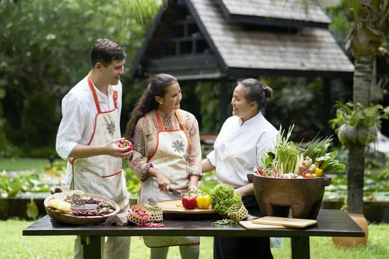 A chef showing fresh vegetables and herbs to two guests in aprons at an outdoor cooking class
