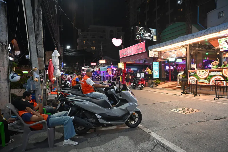 Motorbike taxi riders in orange vests waiting beside parked scooters on a Thai street at night