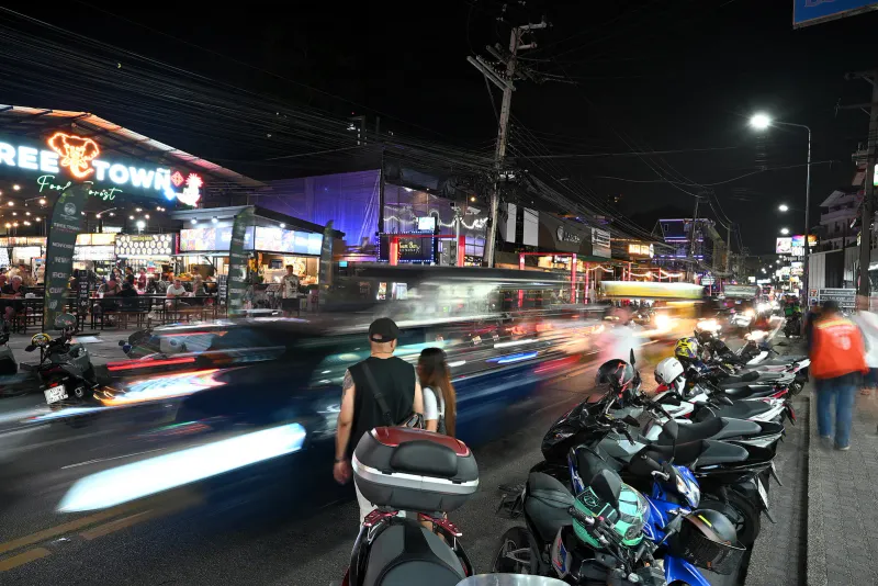 Row of parked scooters and helmets beside a busy night street in a Thai tourist town