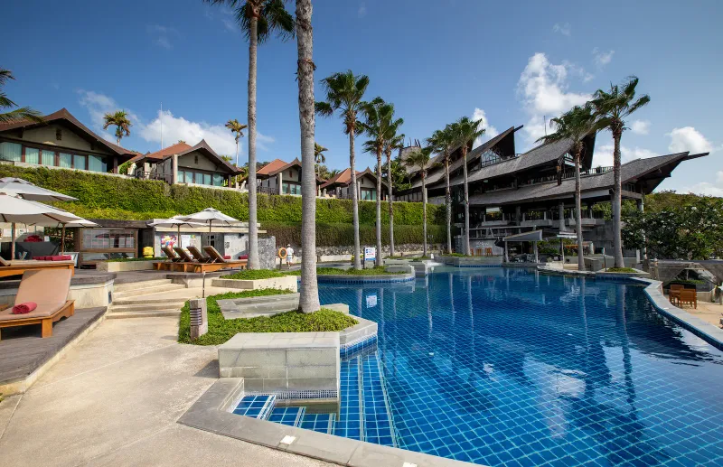 Curved blue infinity pool with sun loungers below the timber roofs of Nora Buri Resort