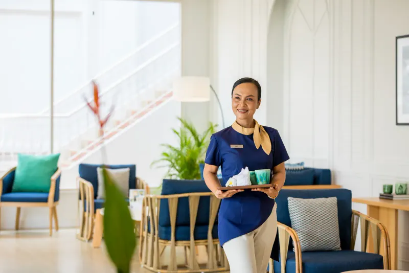 Hotel staff member in a blue uniform holding a tray in a bright lobby lounge at Outrigger Koh Samui