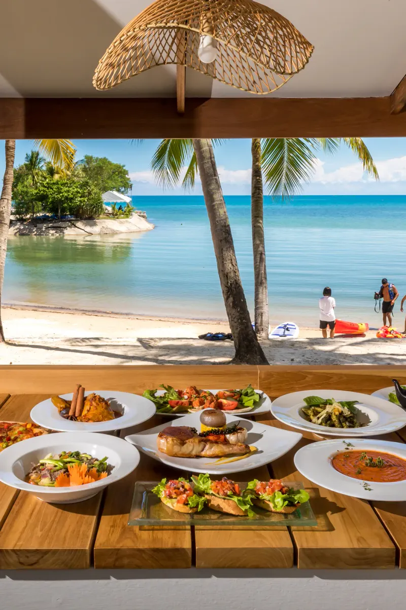 Palm-lined sandy beach with kayaks and a table of Thai dishes from the Outrigger Koh Samui food and drink page