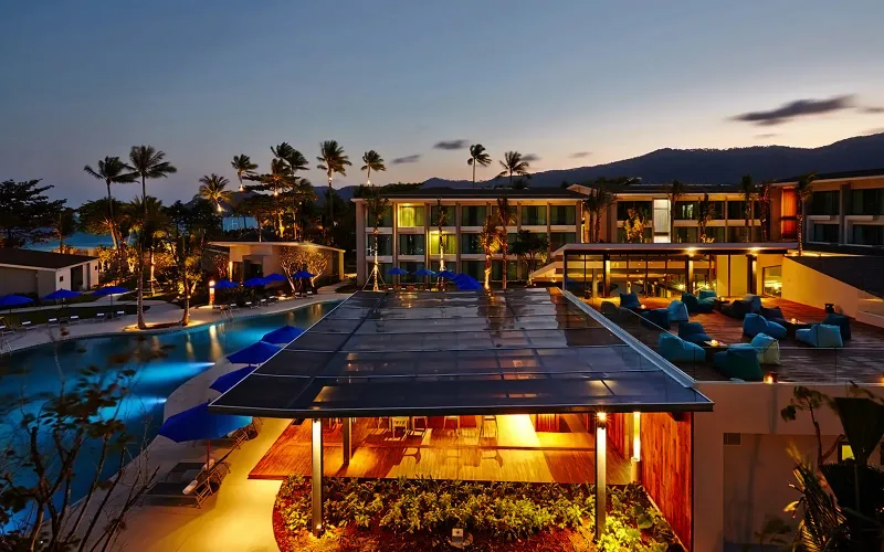 Dusk view of the OZO Chaweng Samui pool, lit bar pavilion and room blocks under palms on Chaweng beach