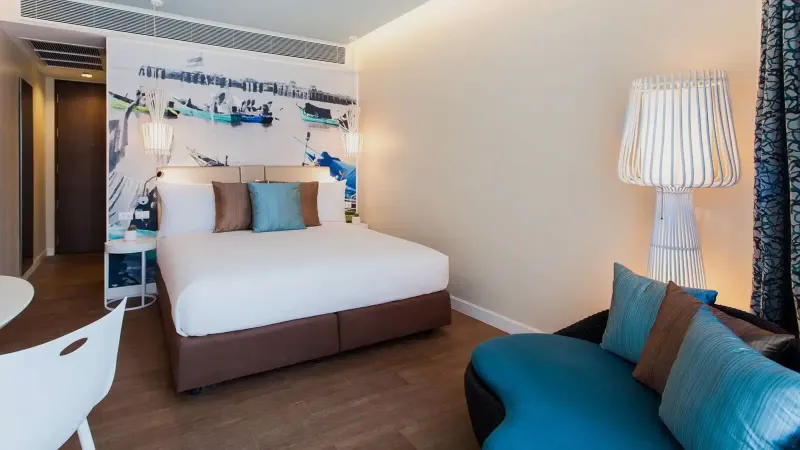 Double bed in an OZO Chaweng Samui room with a fishing boat mural, turquoise cushions and a white lamp