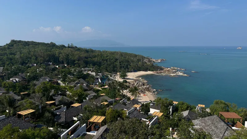 Erhöhter Blick auf die Resortdächer, den kleinen Sandstrand und die felsige Uferlinie der privaten Bucht