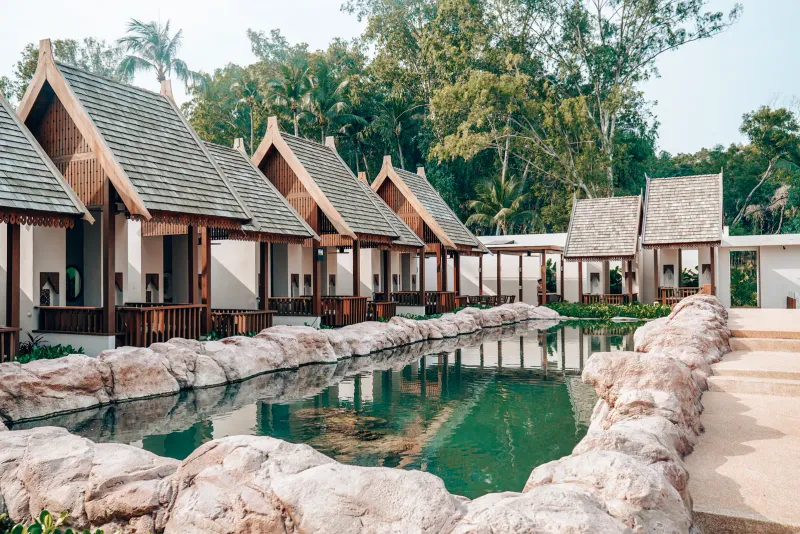 Reihe thailändischer Holzpavillons mit Spitzdächern an einem felsgesäumten Teich im Resortgelände