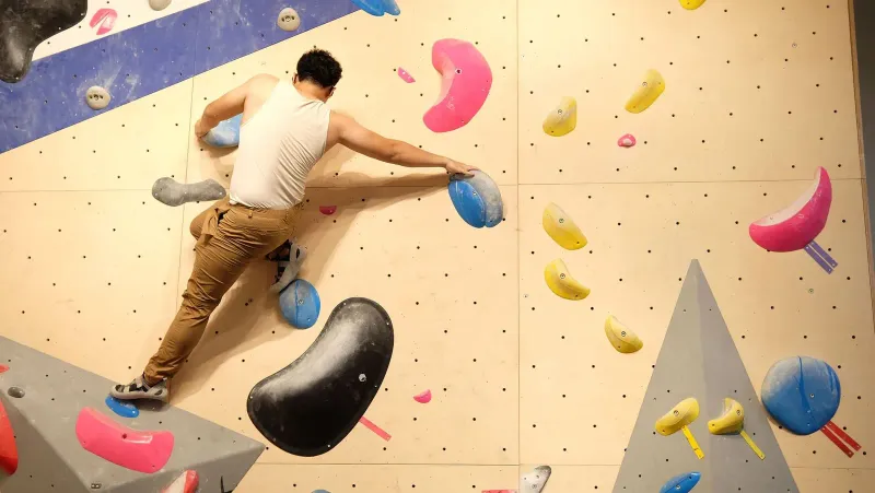 Climber stretching between two holds on a bouldering wall, the indoor training style Samui's own sports park now confirms