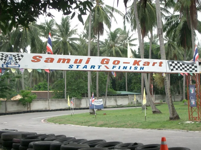 Weathered start/finish banner reading Samui Go-Kart over an empty asphalt track lined with tyre barriers, Bophut