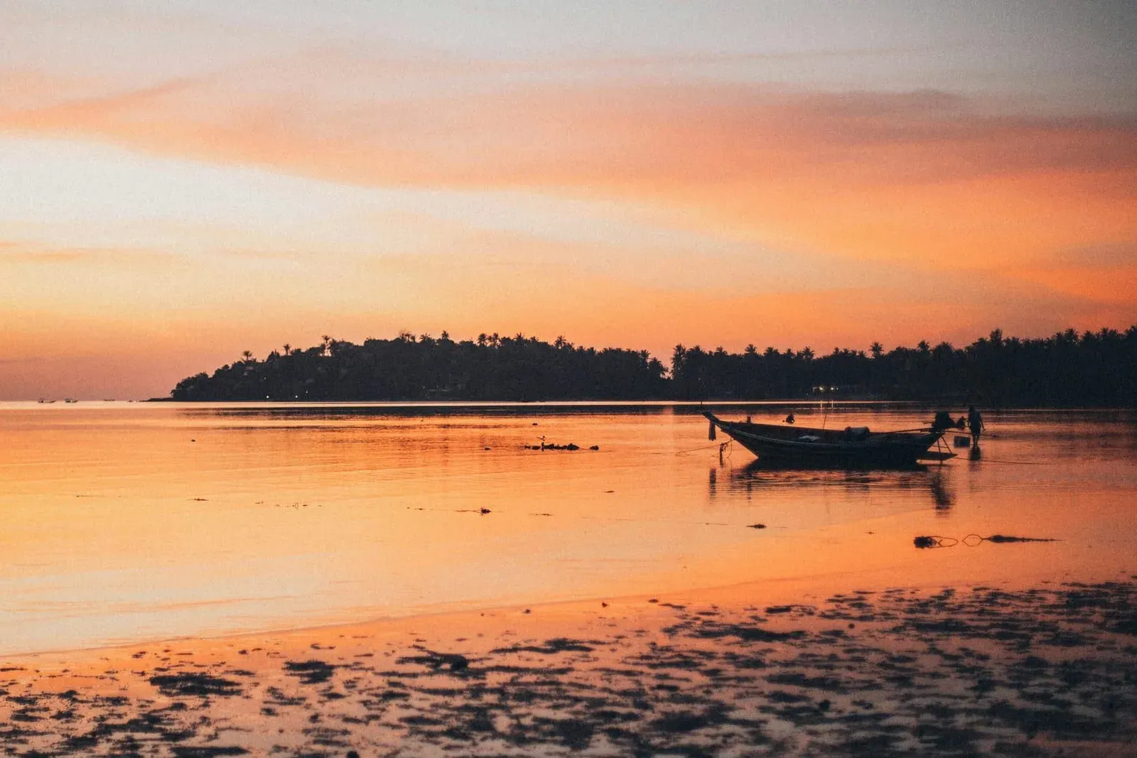 Wooden longtail boat resting in shallow water at low tide off Lipa Noi, a palm-covered headland in silhouette, sky and wet sand glowing orange after sunset