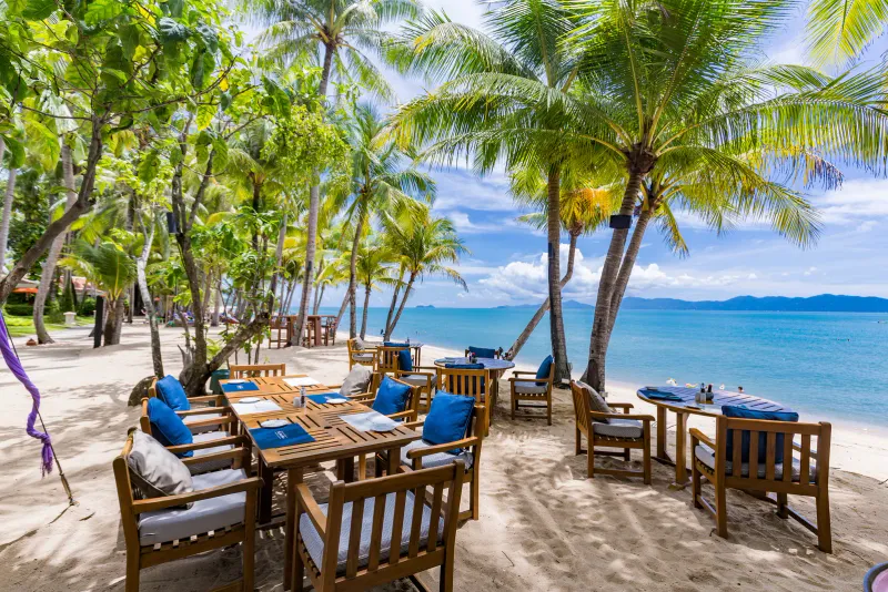 Wooden dining tables set on the sand under coconut palms beside the sea at Santiburi Koh Samui