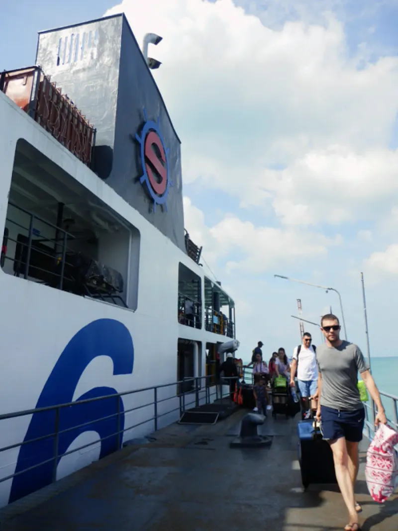 White Seatran ferry with a large S wheel logo as passengers with wheeled suitcases walk along the pier