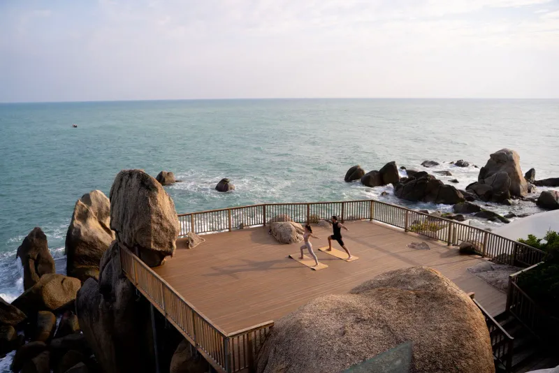 Two people practising yoga on a timber deck built over granite boulders above the sea