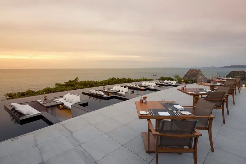 Open-air rooftop restaurant terrace with sunken lounge seats and dining tables facing the sea at sunset