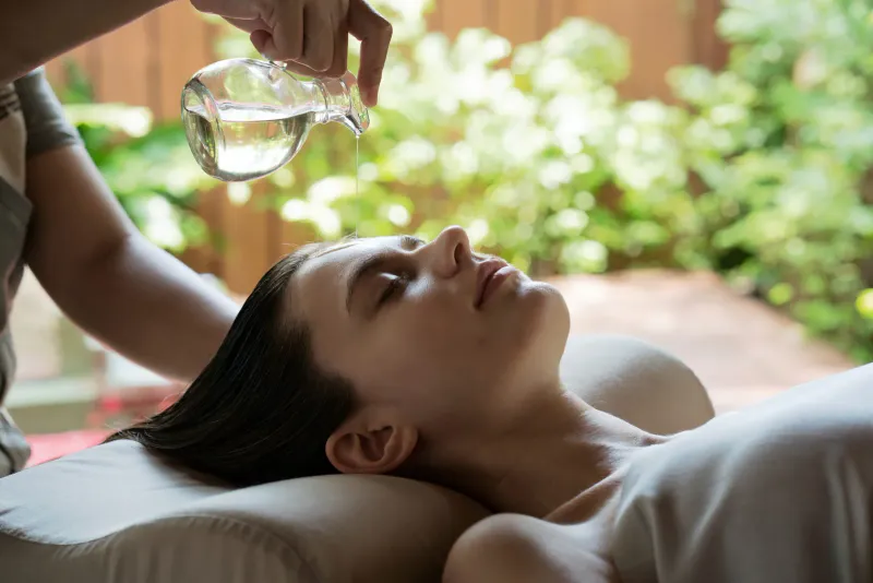 Spa therapist pouring oil over a guest's forehead during a treatment in a garden-facing room