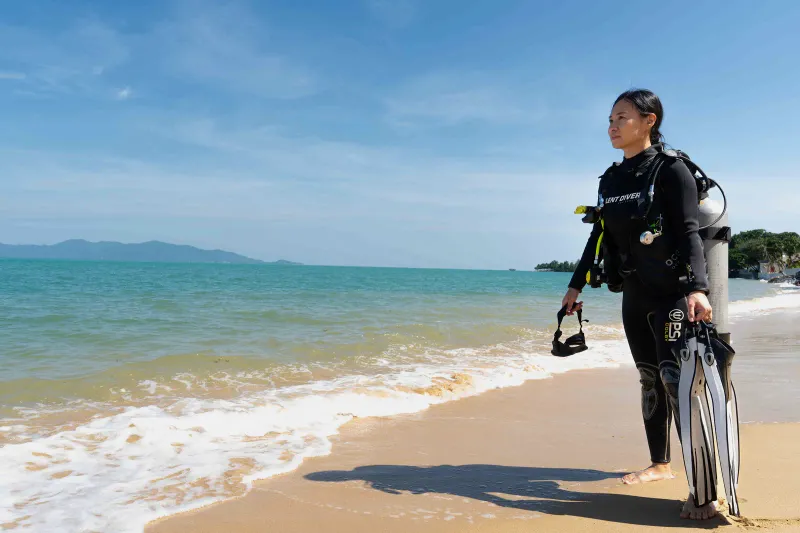 A diver in a wetsuit carrying fins on a sandy beach