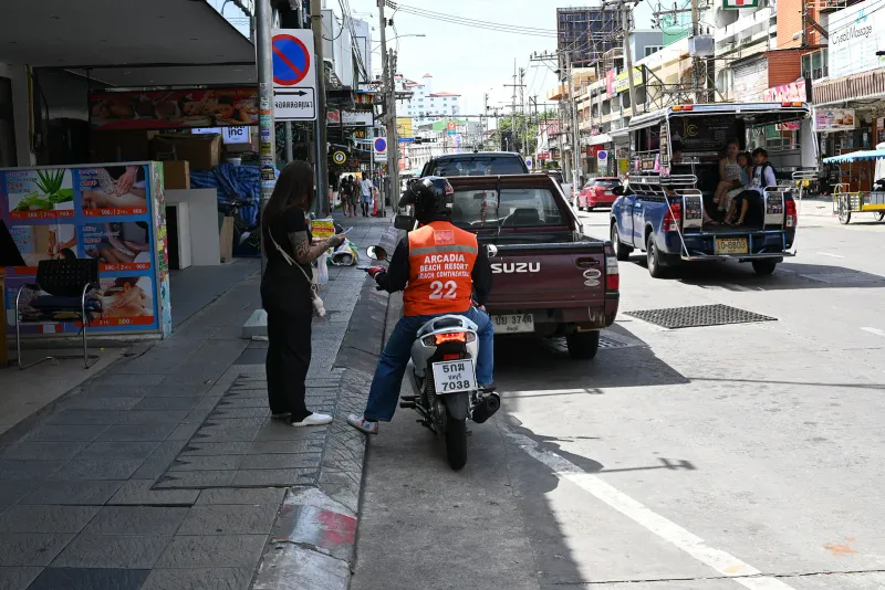 Motorbike taxi rider in an orange numbered vest waiting at a Thai roadside next to a songthaew