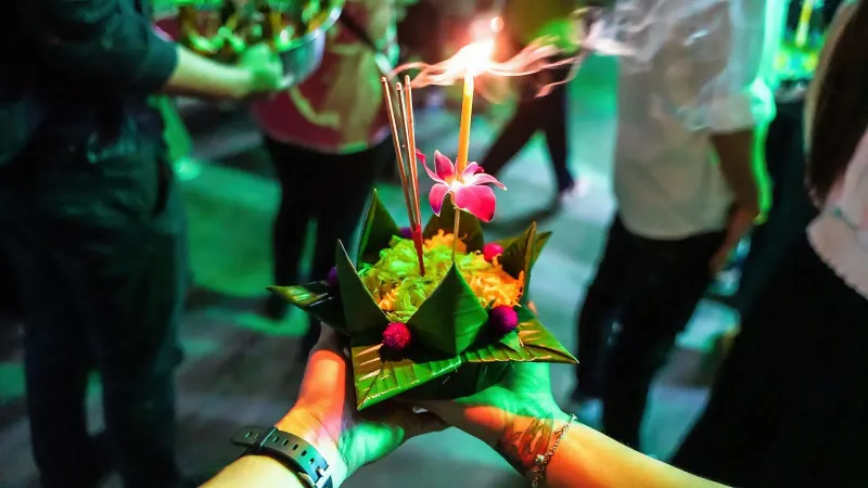 Hands holding a decorated krathong offering with a candle and flowers before setting it on the water