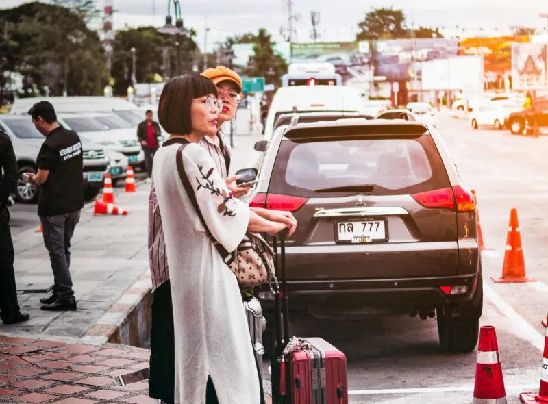 Travellers with luggage waiting beside a parked car with traffic cones on a busy Thai street