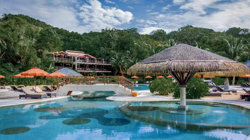 Resort swimming pool with thatched-roof sala and lounge chairs beneath the hillside