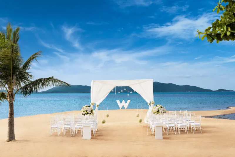 Beach wedding set-up on the sand at W Koh Samui with white chairs, an arch and the island behind