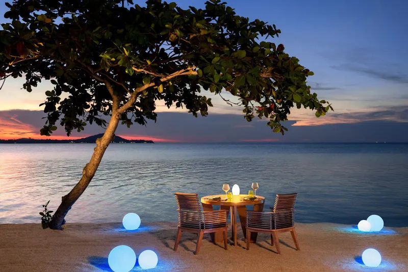 Candlelit table for two on the sand at dusk with glowing spheres and the sea behind