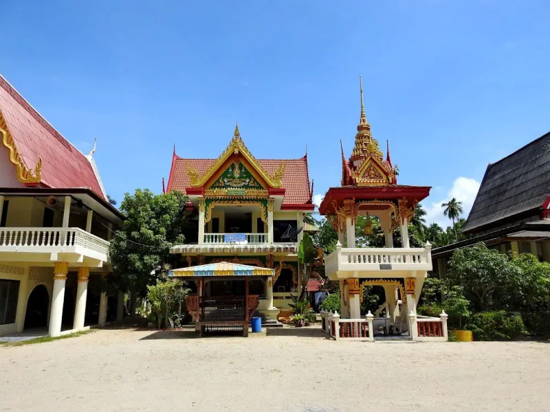 A Thai temple hall and small red-and-gold bell pavilion in a sandy courtyard