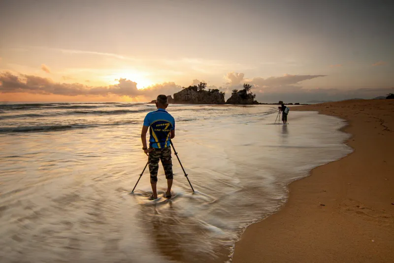 A photographer with a tripod at the water's edge at sunset